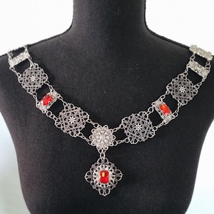 Peut inclure: Collier en filigrane argenté avec des accents de pierres précieuses rouges. Le collier présente un design floral et géométrique complexe, avec un pendentif central et des pierres rouges serties dans des cadres décoratifs. Le collier est présenté sur un mannequin noir.