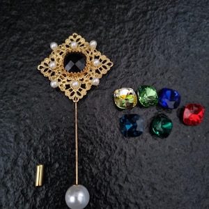 Può includere: Una spilla dorata con una pietra centrale nera e piccole perle. La spilla ha un lungo spillo con una perla all'estremità. Sono presenti anche sei gemme colorate a taglio quadrato: giallo, verde, blu, verde acqua e rosso.