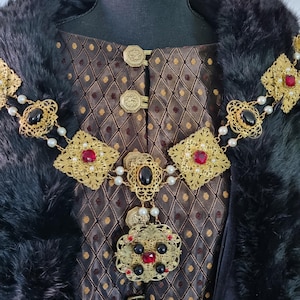 Peut inclure: Un collier de chaîne en or avec des pierres précieuses rouges et noires, porté sur un tissu à motif brun et or avec une bordure en fourrure noire.