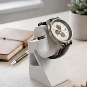 Könnte beinhalten: Ein weißer, S-förmiger Uhrenständer, der eine silberfarbene Chronographen-Armbanduhr mit weißem Zifferblatt und dunkelbraunem Lederarmband hält. Der Ständer hat eine glatte, matte Oberfläche und ein geometrisches Design für einen stabilen Halt.