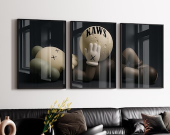 May include: A set of three framed wall art prints featuring a stylized character with a round, moon-like head, tan skin, and gloved hands with X-shaped eyes. The central print displays the text KAWS in bold black letters across the top.