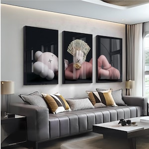 Pode incluir: Três obras de arte emolduradas com figuras de arte pop. Uma mostra uma figura branca semelhante a um urso com olhos cruzados pretos, outra uma figura rosa segurando dinheiro e a terceira uma figura rosa em uma pose relaxada. A arte está pendurada acima de um sofá cinza.