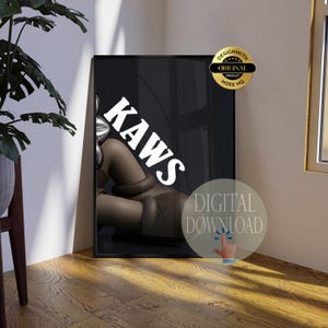 May include: A rectangular black framed wall art print featuring a brown cartoon character with X-shaped eyes and the white text KAWS. The print includes a gold circular seal labeled DESIGN WITH ORIGINAL PRODUCT MZEE HQ and a digital download icon.