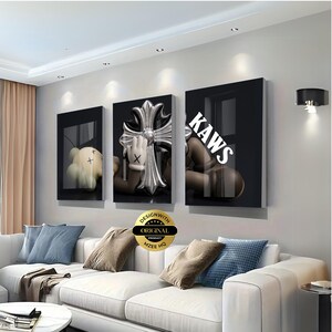 May include: A set of three rectangular wall art panels featuring a black background with a brown cartoon character with X-shaped eyes, a metallic silver cross emblem, and the white text KAWS printed across the right panel in a bold font.