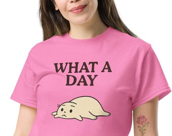 May include: Pink t-shirt with the text "WHAT A DAY" above a cartoon illustration of a sad, light-tan blob. The shirt is made of a soft material and is suitable for casual wear.
