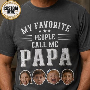 May include: Dark gray t-shirt with the text "MY FAVORITE PEOPLE CALL ME PAPA" in white. The shirt features four cartoon faces below the text. A gold and white circular logo is in the upper left corner.