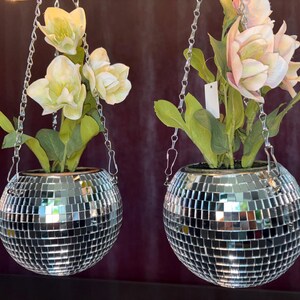 Op de afbeelding: Twee bolvormige hangende plantenbakken bedekt met kleine reflecterende zilveren spiegeltegels. Elke plantenbak hangt aan een zilveren metalen ketting en bevat kunstbloemen met groene bladeren. De linker plantenbak bevat witte bloemen, de rechter roze.
