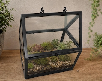 Könnte beinhalten: Ein geometrisches Terrarium mit einem schwarzen Metallrahmen und klaren Glasscheiben. Die Struktur verfügt über einen Klappdeckel mit Verschluss und zwei obere Ringe zum Aufhängen. Im Inneren befinden sich Erde, kleine Kiesel und Sukkulenten.