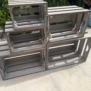 Puede incluir: Un conjunto de cuatro cajas de madera rectangulares con un acabado gris desgastado, apiladas en una disposición escalonada. Cada caja presenta lados y bases de listones con asas recortadas integradas en los extremos, unidas con cuerda fina.