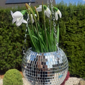Könnte beinhalten: Ein kugelförmiger Disco-Kugel-Pflanztopf, der mit kleinen, reflektierenden silbernen Spiegelkacheln bedeckt ist. Der Topf hängt an einer silbernen Metallkette und enthält künstliches grünes Gras sowie weiße Schneeglöckchen mit grünen Stielen.