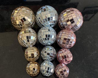 May include: An assortment of disco balls in gold, silver, and rose gold. The spherical ornaments are covered in small, reflective mirrored tiles. The arrangement includes various sizes, creating a visually appealing display for party decorations or home decor.