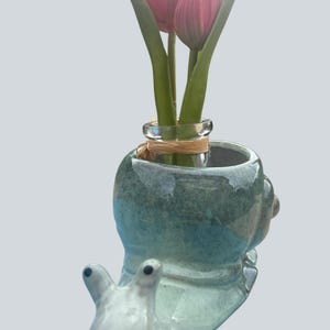 Könnte beinhalten: Ein skurriles Schnecken-Vase aus Keramik in Blau- und Grüntönen, mit zwei rosa Tulpen. Die Schnecke hat ein detailliertes Gesicht und eine kleine Glasvase in ihrem Gehäuse, die mit Bindfaden gebunden ist. Ein einzigartiges Wohnaccessoire.