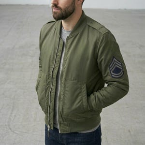 Könnte beinhalten: Olivgrüne Bomberjacke mit silbernem Reißverschluss, zwei Vordertaschen und geripptem Saum und Bündchen. Die Jacke hat einen Aufnäher im Militärstil auf dem linken Ärmel. Getragen über einem grauen T-Shirt und blauen Jeans.