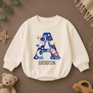May include: A cream-coloured long-sleeve sweatshirt featuring a large letter A with a space-themed graphic including an astronaut, a rocket, a planet, a moon, and stars. The name Anderson is printed in black text below the letter A.