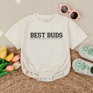 May include: Cream-colored baby onesie with the text "BEST BUDS second generation" in black. The onesie has short sleeves and three snaps at the bottom. Pink sunglasses and baby shoes with smiling faces are also in the image.