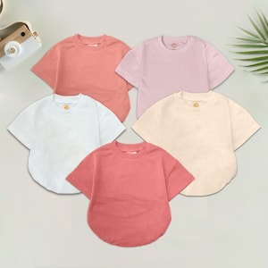 May include: Five short-sleeved t-shirts in various pastel colors: coral, pink, white, cream, and a deeper coral. Each shirt has a round neckline and a relaxed fit. The shirts are arranged in a circular pattern.