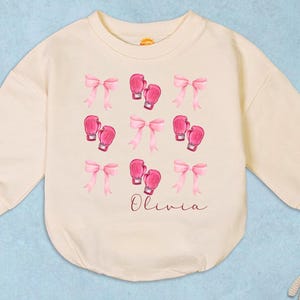 May include: Cream-colored baby bodysuit with long sleeves, adorned with pink bows and boxing gloves. The name "Olivia" is written in cursive script. A charming and playful design for infants.