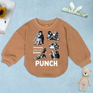 May include: A tan-colored baby sweatshirt with long sleeves. The front features a graphic of several cartoon monkeys in various poses, with the word "PUNCH" in large white letters. The text "Ultimately I have to be brave" is also included.