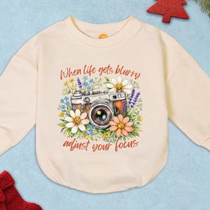 May include: Cream-colored baby onesie with a vintage camera graphic surrounded by flowers. The text "When life gets blurry adjust your focus" is printed above and below the camera. The onesie has long sleeves and a round neckline.