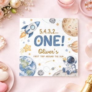 May include: A square paper napkin featuring a space theme with a rocket, a smiling moon, a planet with rings, the Earth, and an astronaut. The text reads 5,4,3,2... ONE! Oliver's FIRST TRIP AROUND THE SUN in blue and gold lettering on a white background.