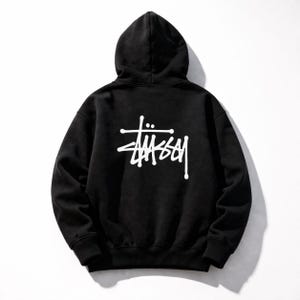 May include: Black hooded sweatshirt with the white "St&uuml;ssy" logo on the back. The hoodie has a drawstring and a kangaroo pocket. The sweatshirt is made of a soft, comfortable material.