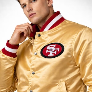 May include: A gold satin bomber jacket with a red and white striped collar and cuffs. The jacket features a team logo patch on the chest. The jacket has a button-up front and two pockets.
