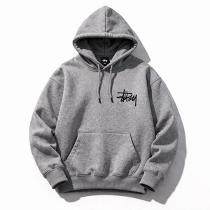 May include: A gray hooded sweatshirt with a front pocket and drawstring. The black logo "St&uuml;ssy" is printed on the front. The sweatshirt is made of a soft material and is suitable for casual wear.