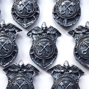 Puede incluir: Diez insignias de metal plateado con una figura de guardián alado sosteniendo una llave. Cada insignia tiene la palabra "GUARDIAN" y un diseño de llaves cruzadas.