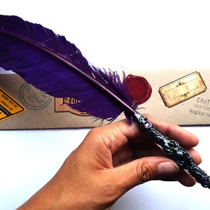 Puede incluir: Una pluma de ave púrpura con un mango de metal plateado y negro. El mango está decorado con un diseño de araña y tiene dos gemas púrpuras. La pluma está apoyada en un paquete de papel marrón con varias etiquetas y sellos.