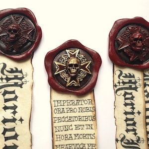 Può includere: Tre segnalibri decorativi con un design a teschio e croce. I segnalibri hanno un sigillo di cera rosso con un emblema a teschio e stella. Il testo sui segnalibri recita "Imperator ora pro nobis peccatoribus nunc et in hora mortis meae voca me."