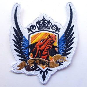 Puede incluir: Parche blanco bordado con un dragón negro y rojo, alas azules y negras, una corona dorada y una pancarta dorada con la palabra "Faust" en ella.