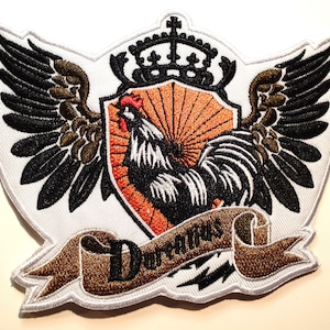 Op de afbeelding: Gestikt patch met een zwart-witte haan, een kroon en vleugels. De haan bevindt zich in het midden van het patch en is omgeven door een bruin lint met het woord "Duratus" erop.