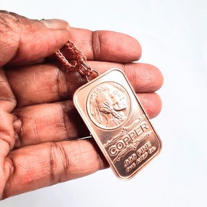 Puede incluir: Un colgante de lingote de cobre rectangular en una cadena de cobre a juego. El lingote presenta un relieve de un perfil nativo americano, la palabra LIBERTY, el texto COPPER, .999 FINE y ONE AVDP OZ. El metal tiene un tono marr&oacute;n rojizo met&aacute;lico.