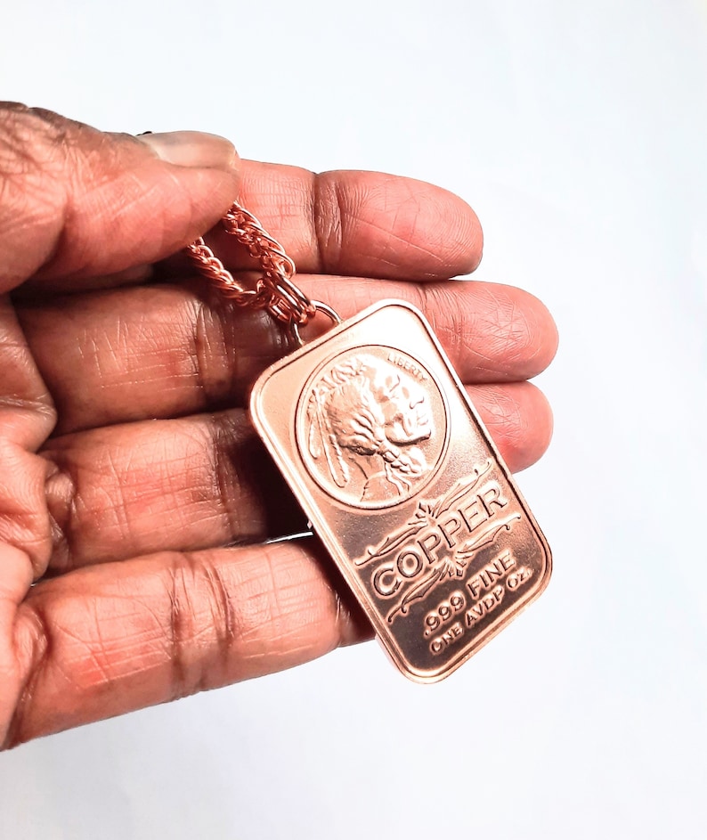 Puede incluir: Un colgante de lingote de cobre rectangular en una cadena de cobre a juego. El lingote presenta un relieve de un perfil nativo americano, la palabra LIBERTY, el texto COPPER, .999 FINE y ONE AVDP OZ. El metal tiene un tono marr&oacute;n rojizo met&aacute;lico.