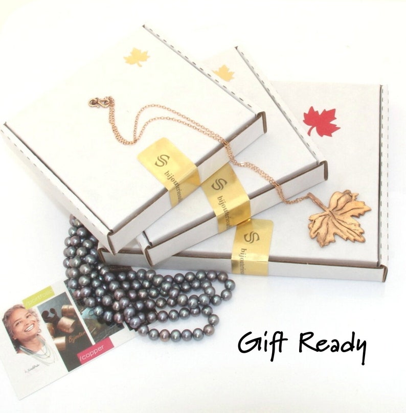 Puede incluir: Tres cajas de cart&oacute;n blanco con pegatinas de logotipo dorado y detalles de hojas de arce, un collar de cadena color cobre con un colgante de hoja de arce texturizado y un collar de perlas gris oscuro. Una tarjeta peque&ntilde;a con texto y Gift Ready.