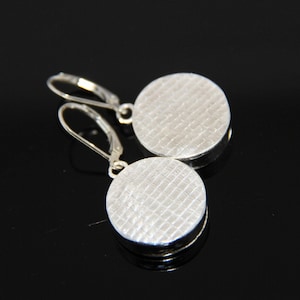 May include: A pair of silver leverback earrings. Each earring features a textured, circular disc. The earrings are displayed against a black background, highlighting their metallic sheen and intricate surface pattern.