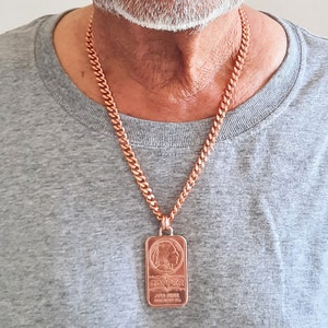 Puede incluir: Un collar de cadena de eslabones de cobre con un colgante rectangular en forma de lingote. El colgante muestra el perfil en relieve de una persona, la palabra COPPER y el texto .999 FINE, ONE TROY OZ.