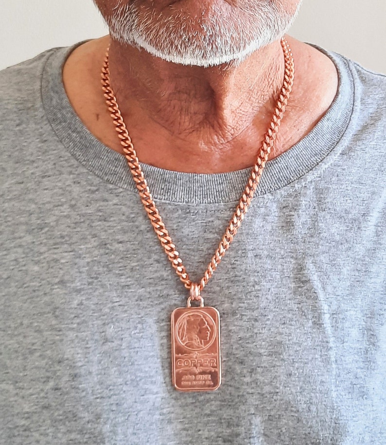 Puede incluir: Un collar de cadena de eslabones de cobre con un colgante rectangular en forma de lingote. El colgante muestra el perfil en relieve de una persona, la palabra COPPER y el texto .999 FINE, ONE TROY OZ.