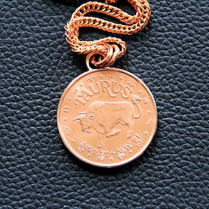 May include: A copper-coloured zodiac pendant necklace with a chain. The pendant features a bull with the word "Taurus" and the dates "April 21 - May 21".