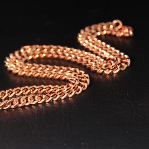 May include: A copper-coloured chain necklace with a curb link design. The chain is displayed on a black surface, with the links reflecting light. The necklace is a jewellery item.