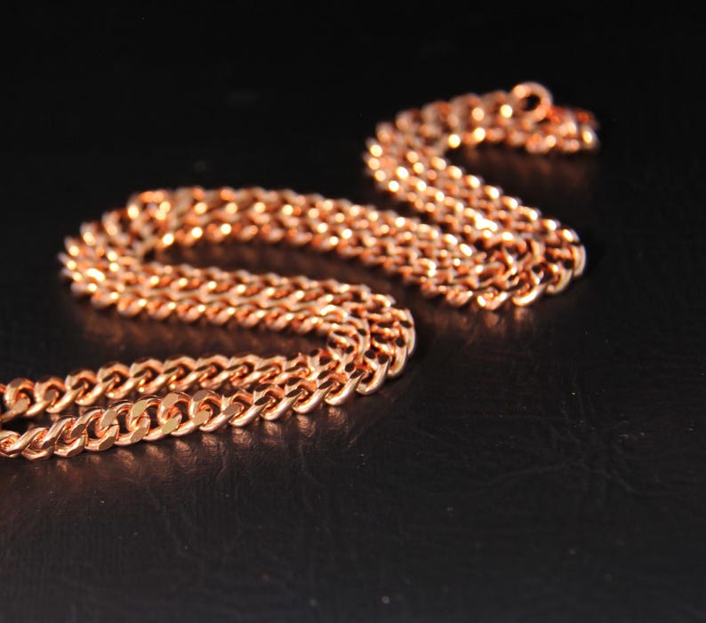 May include: A copper-coloured chain necklace with a curb link design. The chain is displayed on a black surface, with the links reflecting light. The necklace is a jewellery item.