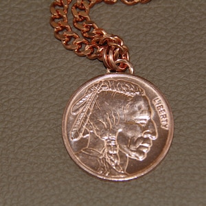 May include: Copper-coloured necklace featuring a round pendant with a Native American profile and the word "LIBERTY." The pendant is attached to a matching copper chain. The necklace is displayed on a textured, neutral-toned surface.