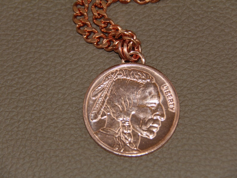 May include: Copper-coloured necklace featuring a round pendant with a Native American profile and the word "LIBERTY." The pendant is attached to a matching copper chain. The necklace is displayed on a textured, neutral-toned surface.