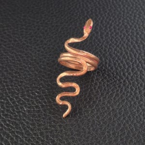 Puede incluir: Un anillo de metal color cobre con forma de serpiente enroscada. El anillo presenta una superficie texturizada y martillada, con dos pequeños acentos circulares de color rosa brillante que sirven como ojos en la cabeza.