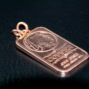 Puede incluir: Un colgante rectangular de cobre con el perfil estampado de una persona con tocado. El texto COPPER y ONE AVDP OZ est&aacute; grabado en la superficie. Una anilla met&aacute;lica circular est&aacute; sujeta a la parte superior de la barra de cobre met&aacute;lico.