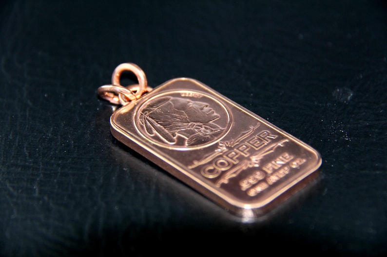 Puede incluir: Un colgante rectangular de cobre con el perfil estampado de una persona con tocado. El texto COPPER y ONE AVDP OZ est&aacute; grabado en la superficie. Una anilla met&aacute;lica circular est&aacute; sujeta a la parte superior de la barra de cobre met&aacute;lico.