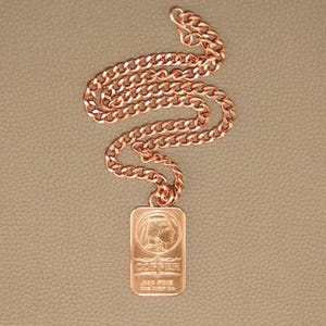 Puede incluir: Un collar de cadena de metal color cobre con un colgante rectangular. El colgante muestra el perfil en relieve de una persona con un tocado, con el texto COPPER, .999 FINE y One AVDP Oz. grabado en la superficie frontal.