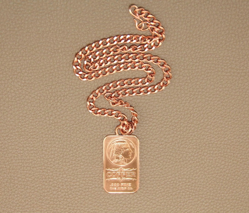 Puede incluir: Un collar de cadena de metal color cobre con un colgante rectangular. El colgante muestra el perfil en relieve de una persona con un tocado, con el texto COPPER, .999 FINE y One AVDP Oz. grabado en la superficie frontal.