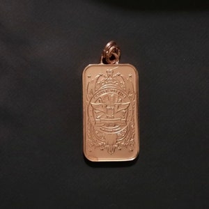 Puede incluir: Un colgante rectangular de cobre con esquinas redondeadas y una anilla en la parte superior. La superficie presenta un dise&ntilde;o en relieve con un emblema alado central, volutas decorativas y el texto COPPER .999 FINE inscrito en el centro.