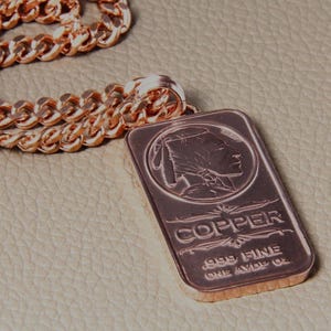 Puede incluir: Un collar de cadena de metal color cobre con un colgante rectangular en forma de lingote. El colgante muestra el perfil en relieve de una figura nativa americana, la palabra COPPER y el texto .999 FINE ONE AVDP OZ.